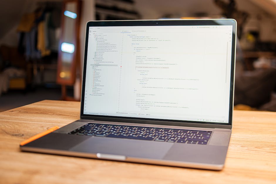 A open laptop placed on a wooden table displays a screen filled with lines of code, set in a cozy indoor environment with warm lighting. The background features blurred household items and furniture, 